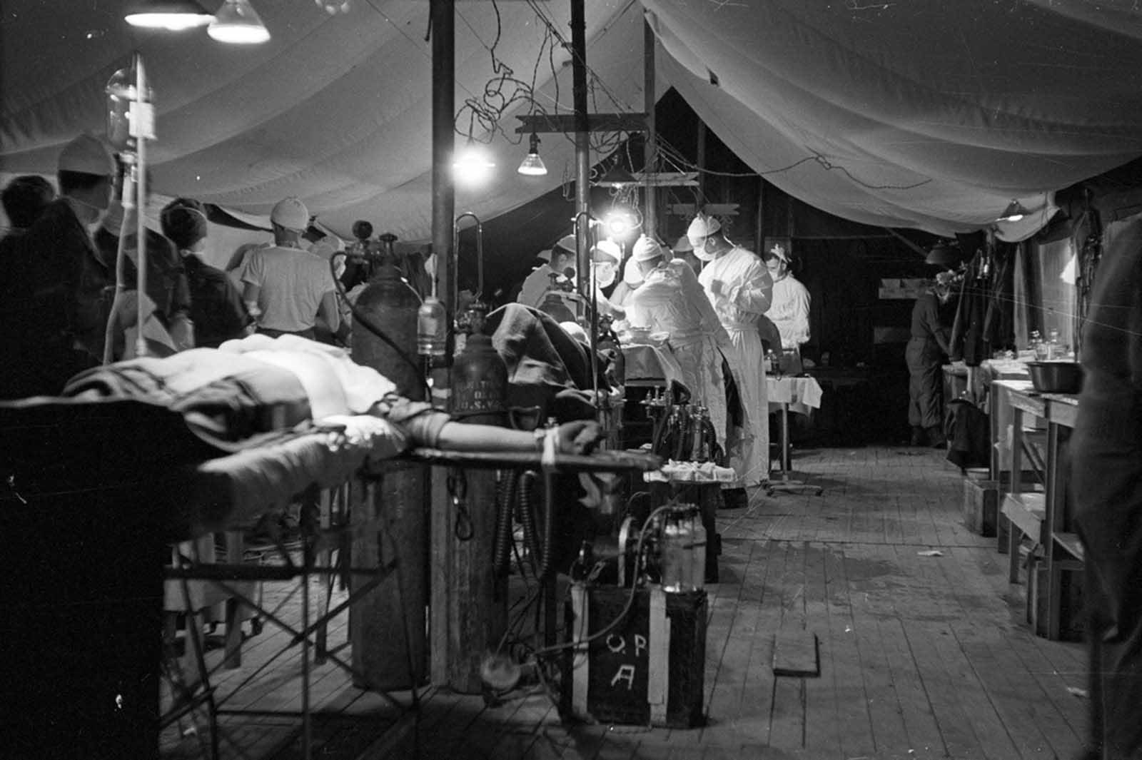 A mobile army surgical hospital somewhere in Korea on October 26, 1951. The patient in the left foreground is receiving blood plasma, while behind him two operations are taking place, one at left and one in the center. Photographer Healy took the photos as he found them. Everyone was so busy that no one had time to pose.